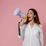 Woman in white shirt holding megaphone against a pink background, confidently speaking.