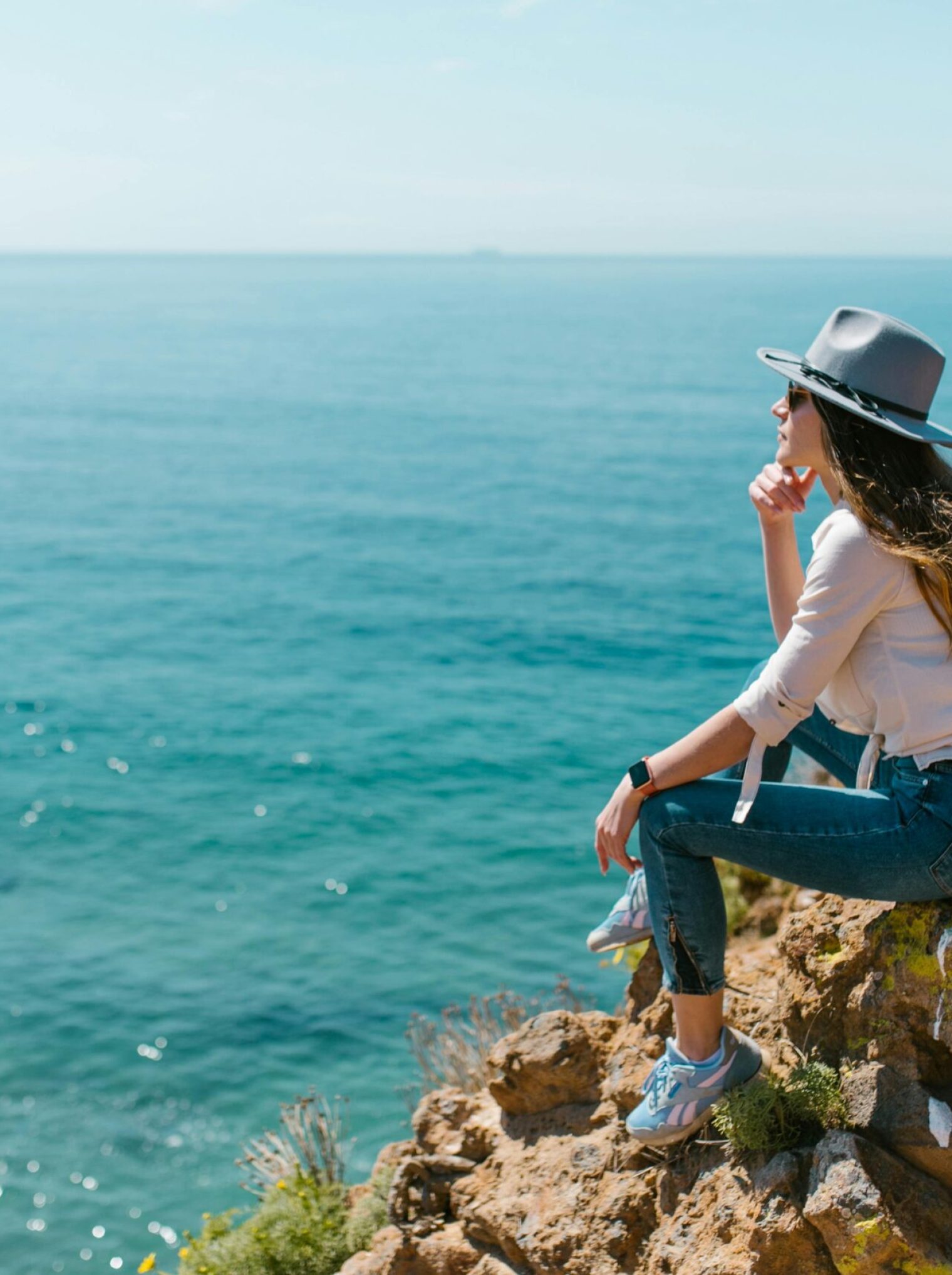 Woman in a hat sitting on a cliff enjoying the ocean view under a clear sky.